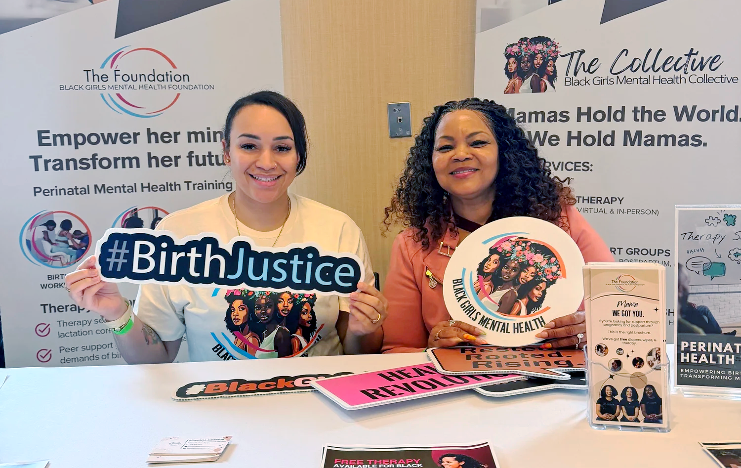 Two women sit at a table with flyers and signs promoting perinatal mental health and birth justice, smiling at the camera, supporting Black Girls Mental Health Collective.
