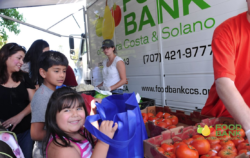 Smiling children and adults gather at an outdoor food bank truck, selecting produce like tomatoes. One girl holds a blue bag, and a staff member stands by crates of vegetables. The Food Bank logo is visible on the truck.