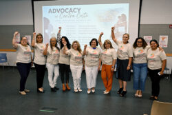 A group of ten people stand in front of a screen that reads Advocacy in Contra Costa. They wear matching shirts and raise their fists, smiling together at the camera in a conference room.