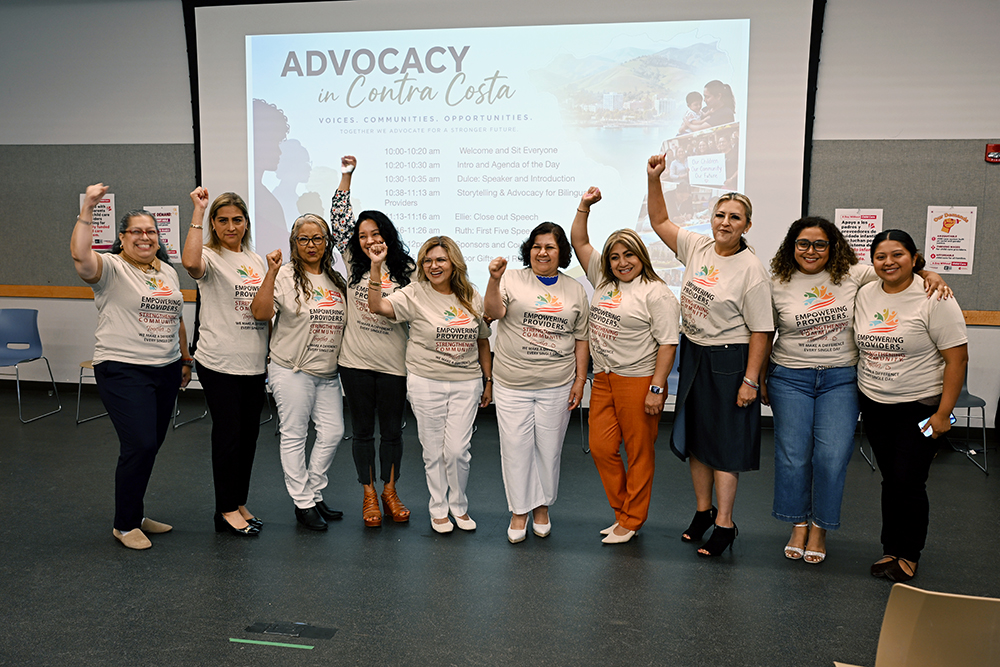 A group of ten people stand in front of a screen that reads Advocacy in Contra Costa. They wear matching shirts and raise their fists, smiling together at the camera in a conference room.