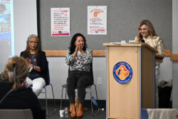 Three women sit and one stands at a podium labeled City of San Pablo. Two posters are visible behind them. The seated women are smiling and clapping while the woman at the podium is speaking.