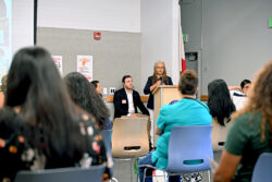 A woman speaks at a podium in a conference room, with several people seated in the audience and panelists sitting beside her.