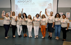 Ten women stand together indoors, smiling and raising their fists. They wear matching t-shirts with a colorful logo and the words Empowering Voices. A presentation slide in the background reads, ADVOCACY in Contra Costa..