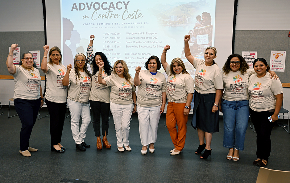Ten women stand together indoors, smiling and raising their fists. They wear matching t-shirts with a colorful logo and the words Empowering Voices. A presentation slide in the background reads, ADVOCACY in Contra Costa..