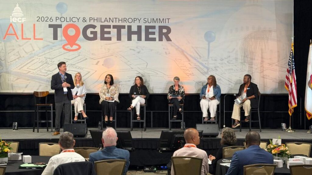 A speaker stands on a stage addressing a seated panel of six people in front of a backdrop for the 2026 Policy & Philanthropy Summit titled All Together.