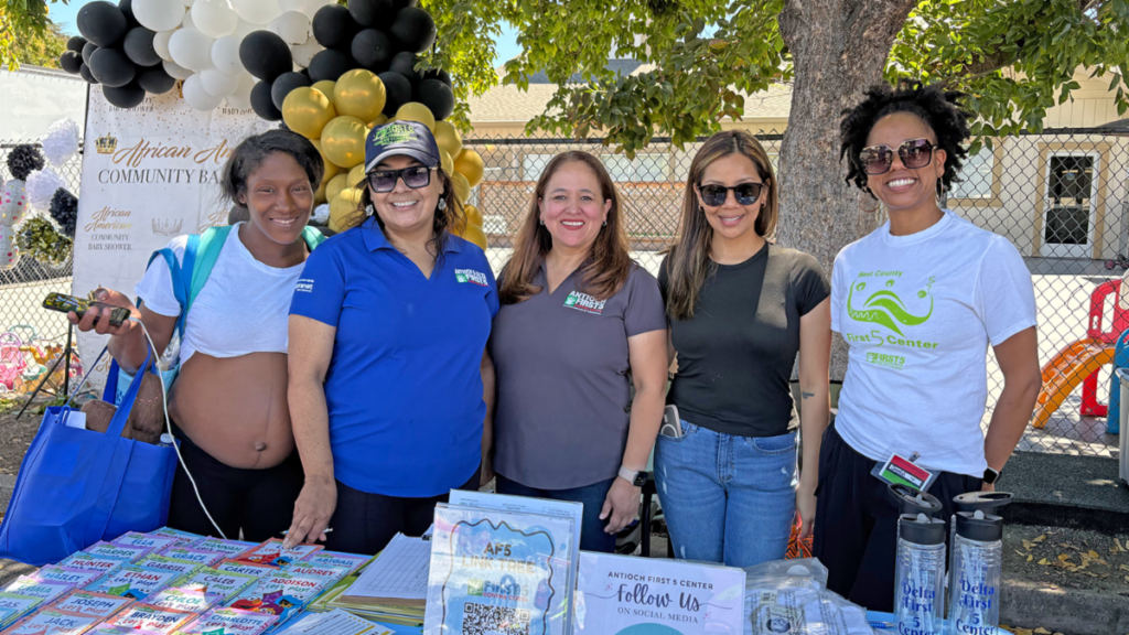 Five women standing behind a table at an outdoor community event, with stacks of books and social media signs on the table in front of them.