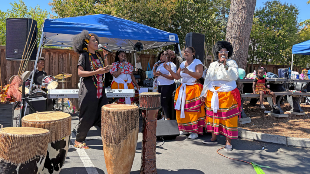 A group of women singing and playing drums on an outdoor stage beneath a blue canopy.