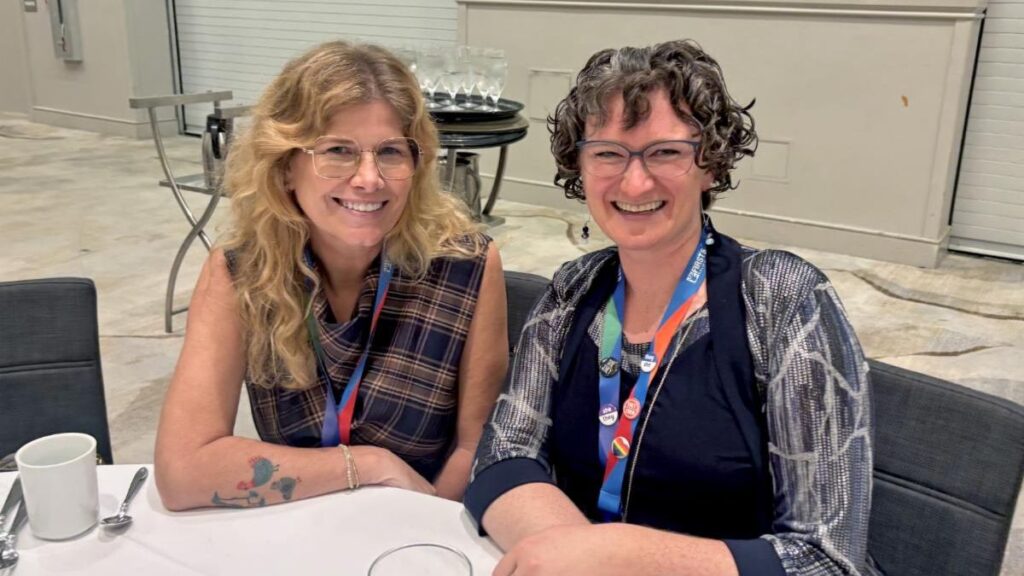 Two women with glasses smile while sitting at a table with name badge lanyards around their necks.