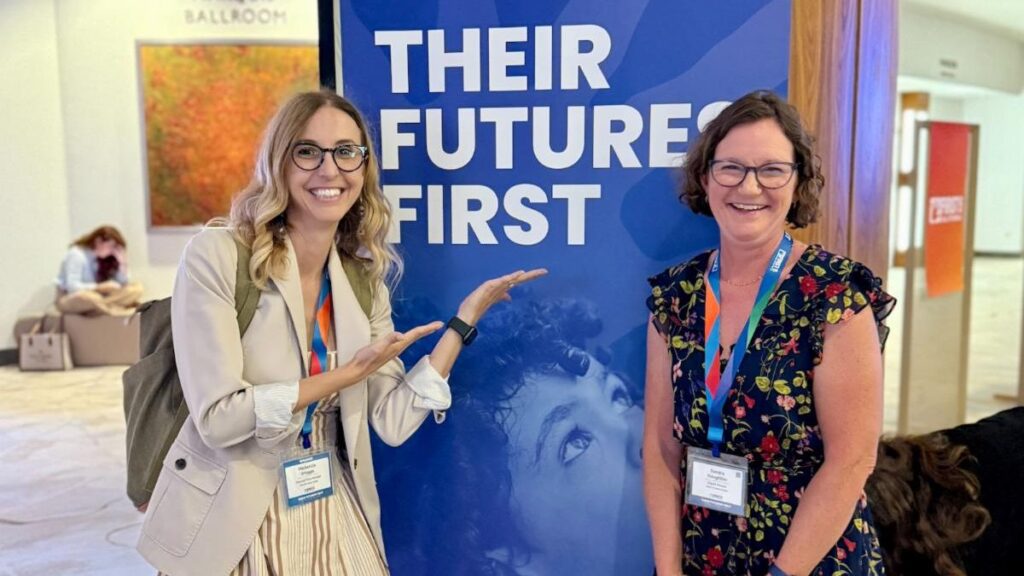 Two smiling women with name tags pose in front of a blue banner that reads, THEIR FUTURE FIRST.